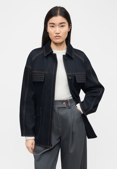 Νεαρή γυναίκα που φοράει σκούρο τζιν μπουφάν με αντίθετη ραφή πάνω από λευκό πουλόβερ και γκρι παντελόνι με ψηλή μέση, στέκεται με το ένα χέρι στην τσέπη.