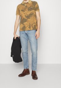 Man med brun kamouflagepolo, ljusblå jeans och mörkbruna läderkängor, som håller en svart jacka i ena handen.