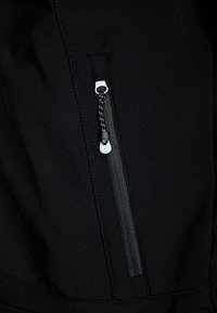 Veste noire avec une poche zippée dotée d'un tirette métallique argenté et d'un tissu texturé. Coutures apparentes le long des bords.