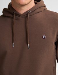 Hoodie marrone realizzato in tessuto morbido, presenta una tasca a canguro frontale, cappuccio con stringhe e puntali in metallo, e un piccolo logo ricamato sul petto.