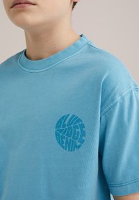 Jasnoniebieska koszulka z krótkim rękawem wykonana z bawełny, z okrągłym nadrukiem graficznym "BLUE RIDGE DENIM" na lewej klatce piersiowej.