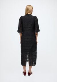 Juoda, tekstūruota midi suknelė su pūstomis rankovėmis, skeltu kraštu ir prigludusia siluetu. Audinys atrodo lengvas ir šiek tiek permatomas.