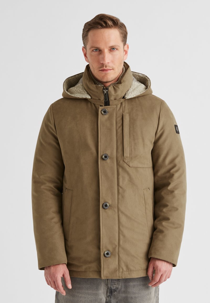 Mann in eine hellbraune Winterjacke mit Kapuze, Knöpfen und Reißverschluss, der vor einem schlichten weißen Hintergrund steht.