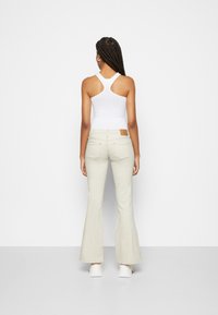 Top de malha branca com alças de nadador, combinado com calças flare bejas claras com bolsos traseiros e um retalho de pele de cor castanha na cintura.