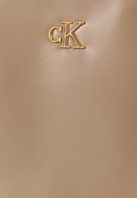 Barna műbőr felület, amelyen egy arany fémes "cK" logó található. Sima textúra enyhe fénylő hatással, minimalista dizájn.