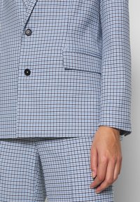 Gros plan sur une veste et un pantalon à motif pied-de-poule bleu et blanc avec des boutons noirs et une main reposant sur le côté.