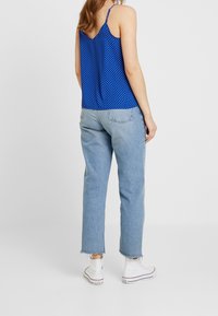 Žena v modré tílkové blůze s puntíky, světlých modrých rovně střižených džínách s třepenými lemy a bílých teniskách na vysoké podrážce, stojí na bílém podlaze.