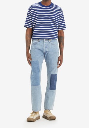 Niebiesko-biała pasiasta koszulka z krótkim rękawem zestawiona z jasnoniebieskimi dżinsami z łatami w różnych fakturach i odcieniach denimu.