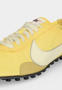 Κίτρινο αθλητικό παπούτσι Nike με λευκά κορδόνια, λογότυπο Swoosh σε λευκό χρώμα και μαύρη υφασμάτινη σόλα, από κοντινή πλευρική γωνία.