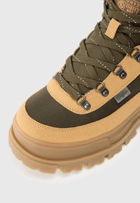 Botte de randonnée verte et beige avec des matériaux en daim et en nylon, semelle en caoutchouc épaisse, système de laçage complexe et accents en métal.