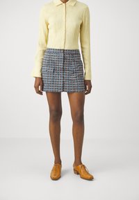 Camisa amarilla de manga larga con textura y cuello, combinada con una falda plisada azul con patrón. Zapatos beige con cordones completan el conjunto.