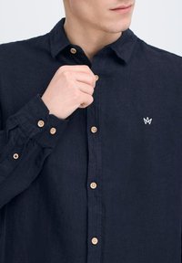 Camicia in lino blu navy con colletto, caratterizzata da bottoni beige e un logo ricamato bianco sul lato sinistro del petto. Tessuto testurizzato.
