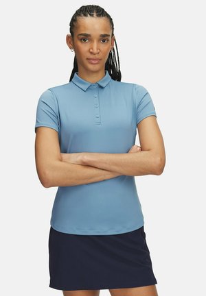 Lyseblåt poloshirt med en blød tekstur, korte ærmer og en knaplukning. Parret med en mørkeblå nederdel.