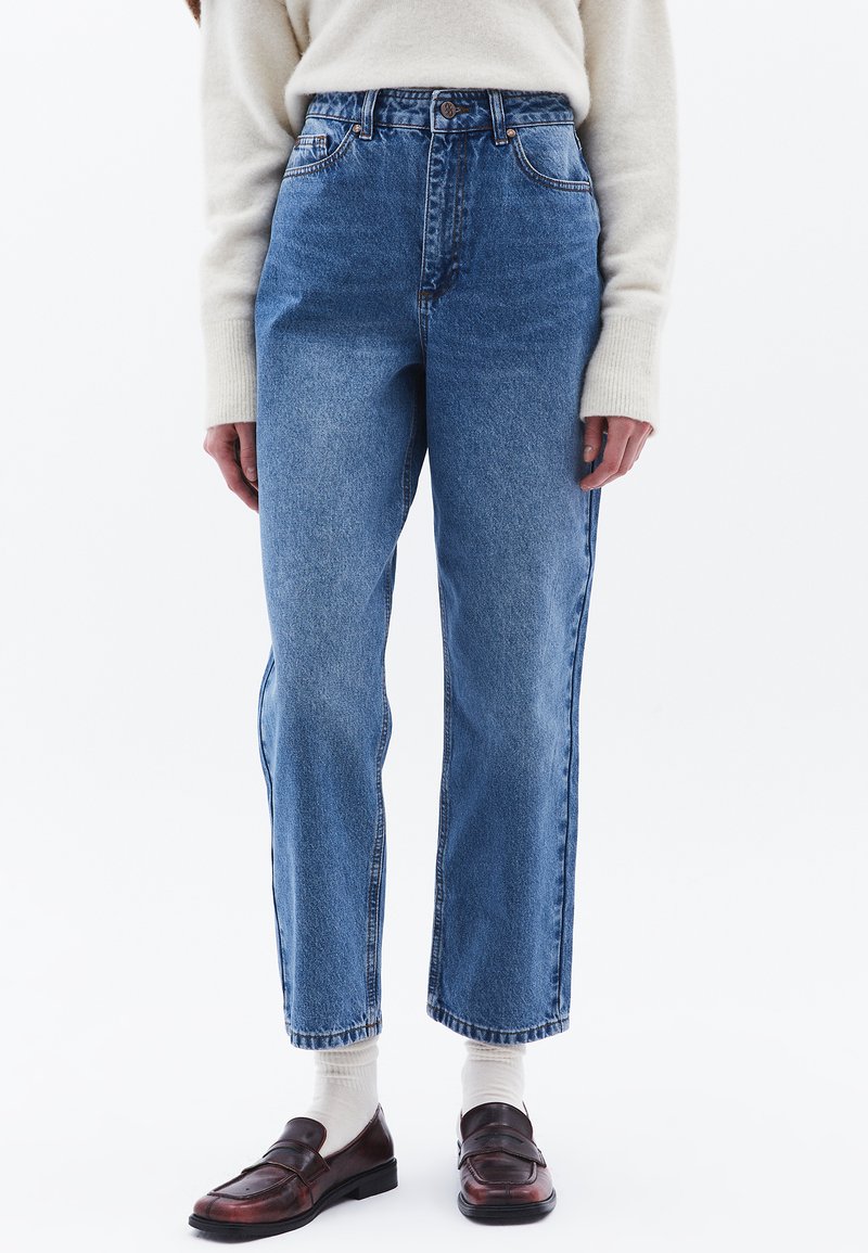 Högmidjade blå denimjeans med rak passform, fem fickor och subtila blekningar. Bärs med bruna läderskor och vita strumpor.