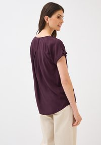 Blouse violette à manches courtes avec un col rond, en tissu doux, et de petits détails noués sur les manches, associée à un pantalon large beige clair.