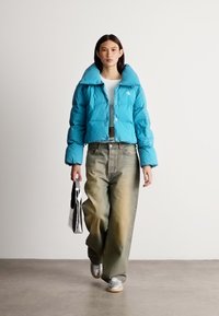 Svijetloplavi puffer kaput s visokim ovratnikom, uparen s izblijedjelim širokim trapericama i srebrnim tenisicama. Model drži crno-bijelu torbu.