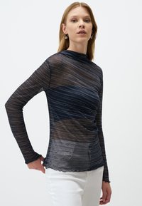 Top blu navy a maniche lunghe con strisce argento orizzontali, colletto alto, texture trasparente e orlo ondulato. Abbinato a pantaloni bianchi.