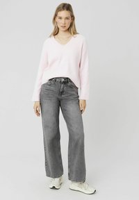 Un pull rose clair à col en V, à la texture douce, associé à un jean large gris taille haute. Des baskets blanches et des colliers superposés sont visibles.
