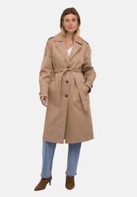 Cappotto beige in trench con cintura, grandi bottoni e spalline; realizzato in tessuto liscio; presenta maniche lunghe e design fino al ginocchio.