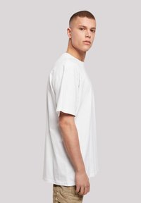 Weißes Baumwoll-T-Shirt mit kurzen Ärmeln, lockerem Schnitt und Rundhalsausschnitt, seitlich an einem Model vor einem schlichten Hintergrund präsentiert.