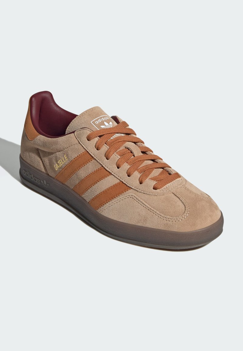 Beżowe zamszowe sneakersy z brązowymi akcentami i trzema paskami z boku. Posiadają bordową podszewkę, okrągły czubek oraz teksturowaną gumową podeszwę.