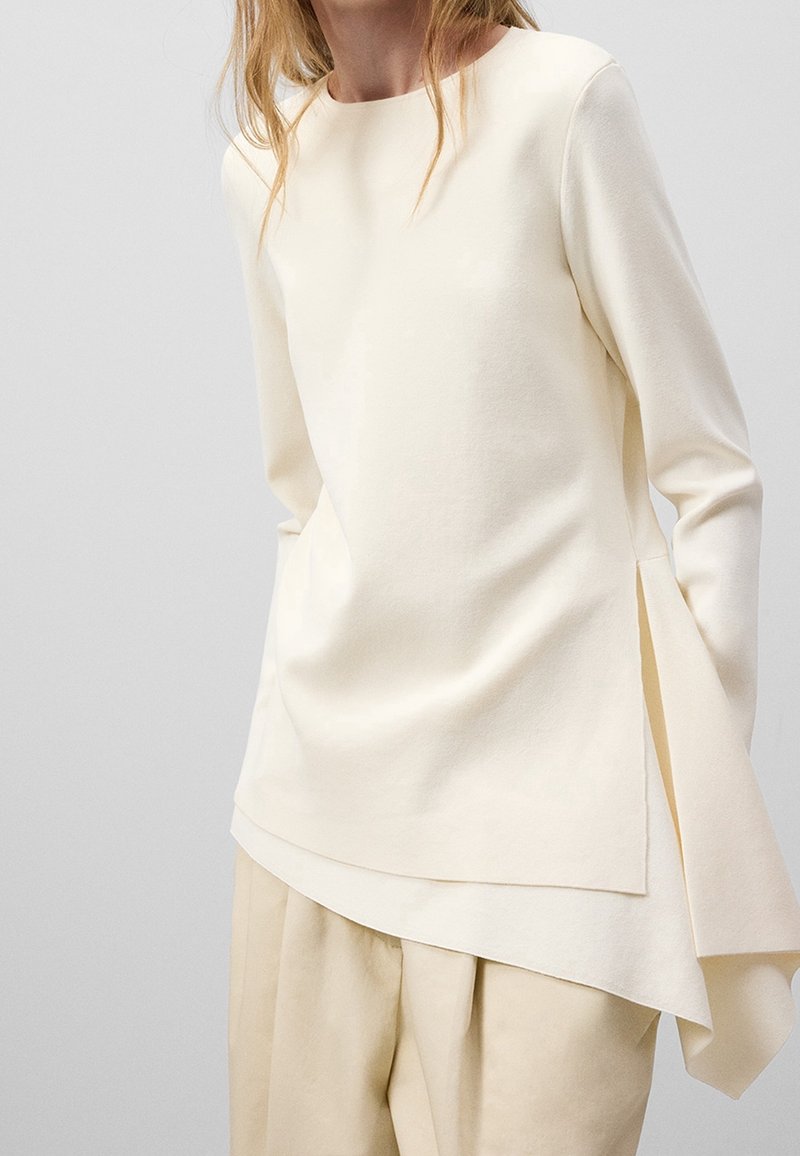 Femme portant un chemisier crème asymétrique à manches longues avec des fentes latérales, associé à un pantalon beige ample assorti sur un fond clair.