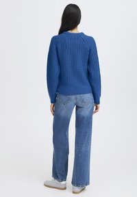 Pull en maille bleue avec un motif texturé, associé à un jean bleu clair ample et des baskets grises. Vue de dos.