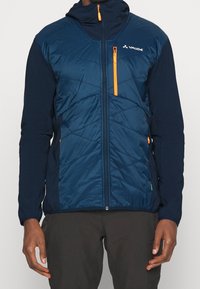 Man som bär en marinblå och mörkblå vadderad jacka med orange dragkedjeakenter och hög krage, tillsammans med mörka byxor.