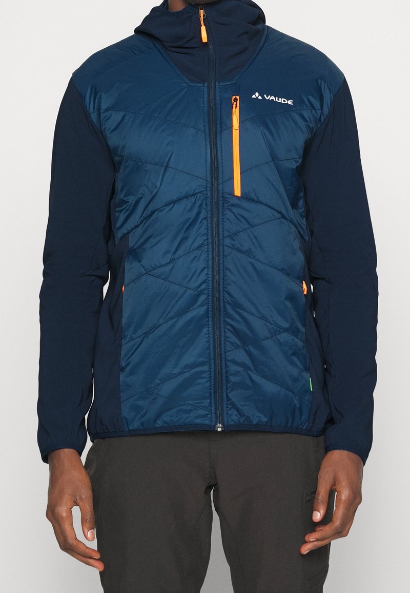 Man som bär en marinblå och mörkblå vadderad jacka med orange dragkedjeakenter och hög krage, tillsammans med mörka byxor.