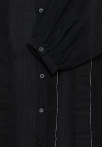 Chemise en tissu noir texturé avec des boutons sur le devant, une manche bouffante repliée et des détails verticaux cousus avec du fil blanc d'un côté.
