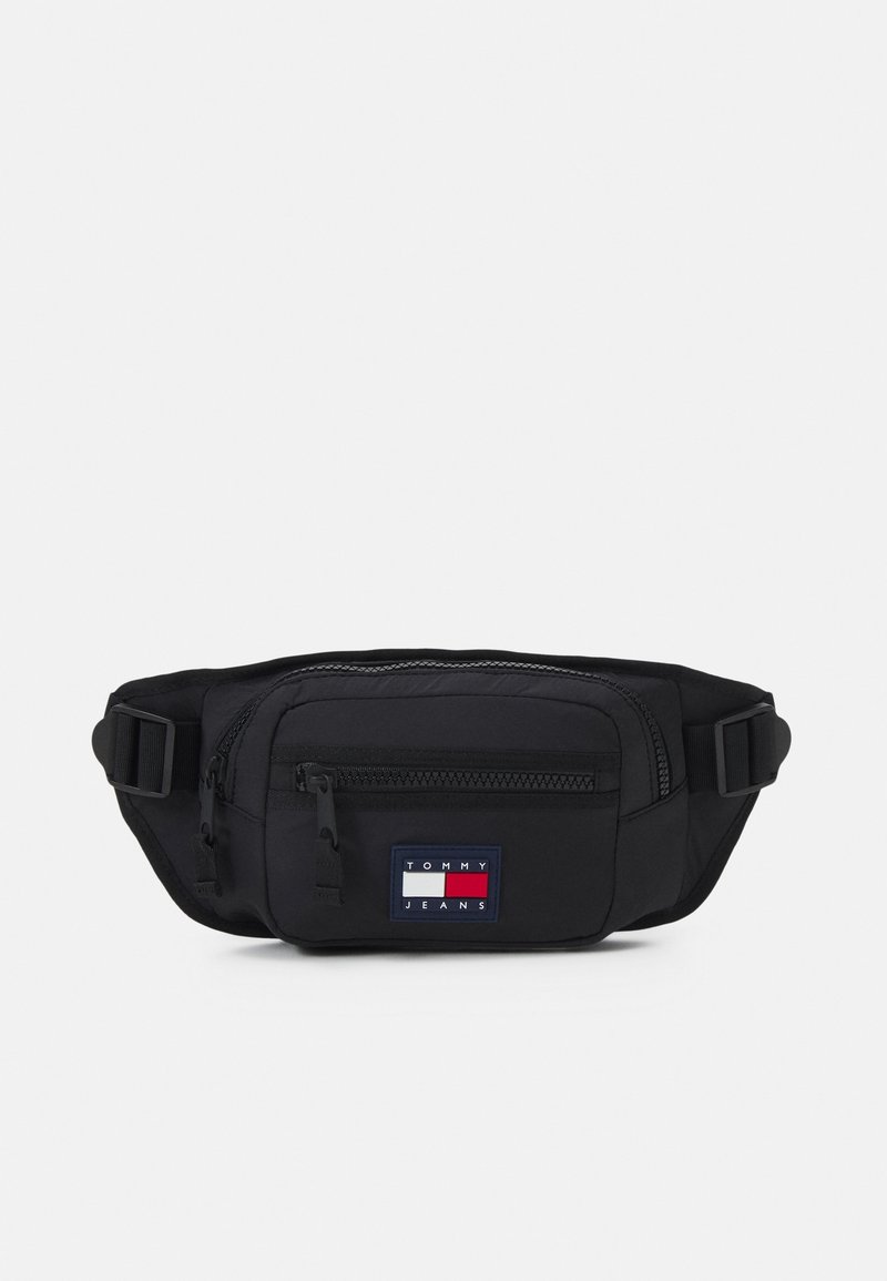 Fanny pack en tissu noir avec un compartiment zippé à l'avant, une sangle ajustable et un patch logo rectangulaire aux couleurs rouge, blanc et bleu.
