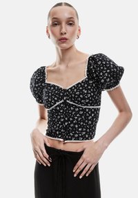 Sort blomster crop-top med puffærmer, udstyret med hvidt blondedetalje og knaplukning. Kombineret med høj talje sorte bukser.