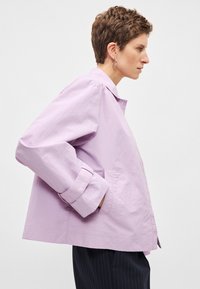 Ανοιχτό μωβ μπουφάν με άνετη εφαρμογή, διπλωμένα μανίκια και πλαϊνές τσέπες· κατασκευασμένο από υφασμάτινη υφή με απλό γιακά.