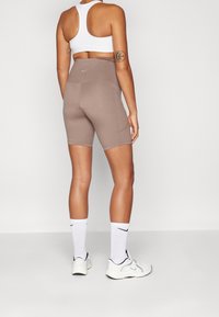 Frau steht und trägt einen weißen Sport-BH, taupefarbene, hoch taillierte Radlerhosen, weiße Nike-Socken bis zur Mitte der Wade und weiße Turnschuhe vor einem schlichten Hintergrund.