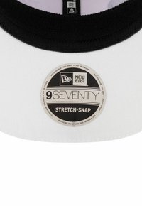 Casquette de baseball blanche avec une visière noire, portant une étiquette ronde qui indique "NEW ERA 9SEVENTY STRETCH-SNAP". Un tissu texturé accentue le design.