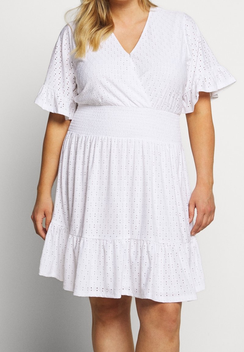 Femme portant une robe blanche en broderie anglaise avec des manches courtes à volants, un col en V croisé, une taille smockée et une jupe à volants qui s'arrête au-dessus des genoux.