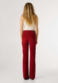 Pantaloni a palazzo in velluto a coste rossi con vita alta, due tasche posteriori e un tessuto texture. Il modello indossa un semplice top bianco senza maniche.