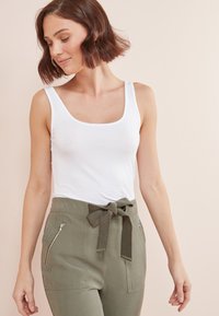Top blanco sin mangas combinado con pantalones verde oliva que presentan una cintura atada y bolsillos con cremallera lateral. Tela suave y diseño ajustado.