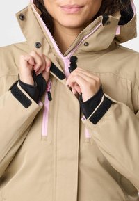 Person, der spænder beige jakke med lyserød lynlås og sorte velcro-stropper på håndleddene, viser delvist ansigt og hænder.