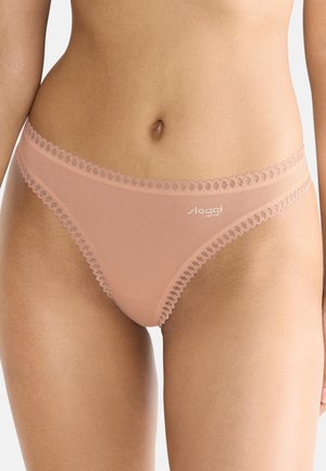 Gros plan sur le torse d'une femme portant un slip rose clair orné de dentelle avec le nom de la marque "sloggi" à l'avant.