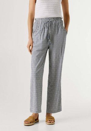 Femme portant un pantalon à rayures verticales bleues et blanches avec un cordon à pompons, un haut sans manches blanc et des sandales marron à enfiler.