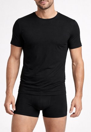 Uomo che indossa una t-shirt aderente nera a maniche corte e boxer abbinati neri davanti a uno sfondo bianco uniforme.