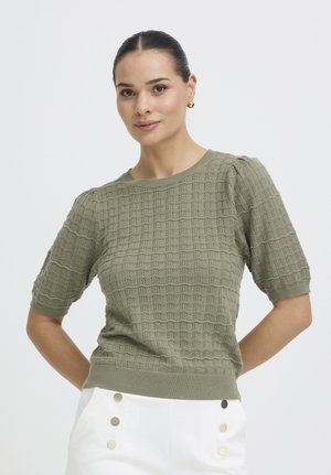 Žena s vlasmi stiahnutými dozadu, má na sebe zlaté náušnice, olivovo zelený textúrovaný sveter s krátkym rukávom a biele nohavice so zlatými gombíkmi, pózuje na bielom pozadí.