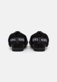 Sandales en fausse fourrure noire avec une large sangle arborant un texte de marque répété. Semelle plate avec une texture douce et un design arrondi.