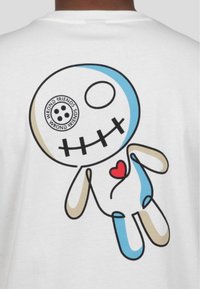 Camiseta blanca con un colorido personaje de dibujos animados con un diseño en azul y beige, cara sonriente, boca negra cosida y un corazón rojo.