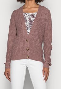 Kvinna som bär en mauve stickad kavaj med tre knappar över en blommig topp, i kombination med vita byxor, som står mot en enhetlig bakgrund.