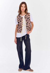 Žena v bílé tričku, leopardím vestě se žlutými tkaničkami, tmavých širokých džínách a hnědých volných botách, stojící s jednou rukou v kapse.