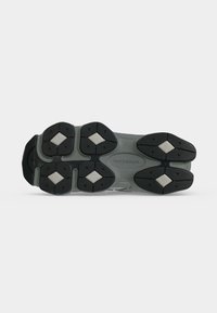 Η σόλα ενός γκρι και μαύρου παπουτσιού New Balance με μοτίβα πέλματος και λευκές περιοχές λαβής σε σχήμα διαμαντιού σε λευκό φόντο.