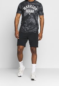 Černé grafické tričko s bílým textem "WARRIOR MANA" a vzory listů, v kombinaci s černými šortkami a bílými teniskami, doplněné černobílými ponožkami.