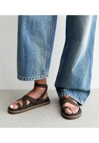 Sandali piatti marroni con più cinturini incrociati, cinturino alla caviglia e suola in gomma, abbinati a jeans a gamba larga. Le scarpe presentano una texture liscia.